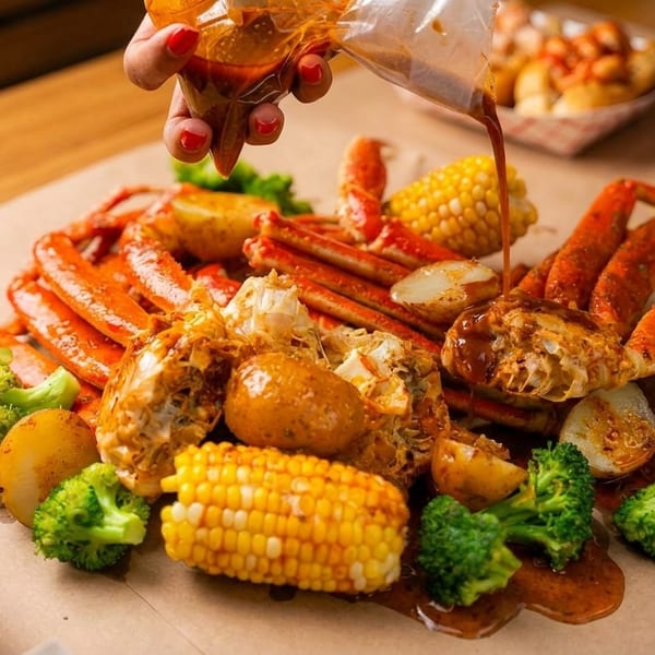 A hand pours sauce over a seafood boil with crab, corn, potatoes, and broccoli on brown paper.