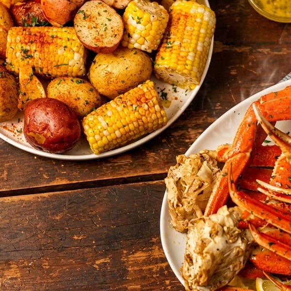 Plates of steamed crab legs, corn on the cob, and potatoes on a rustic wooden table.